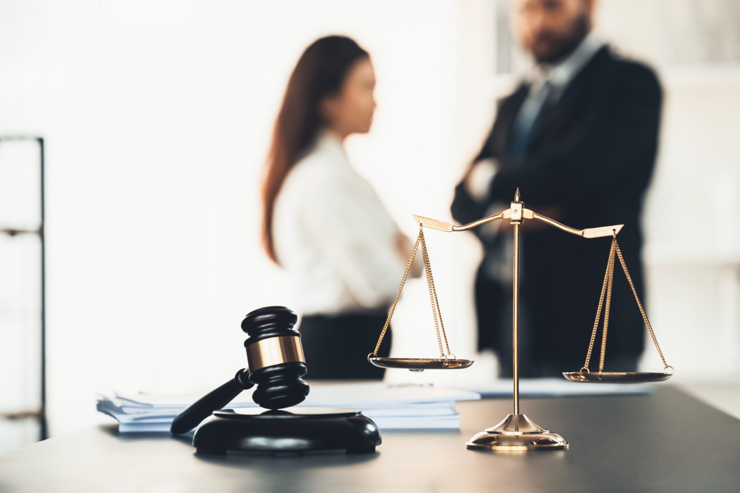 A judge's gavel and scales of justice on a desk, with two blurred professionals in the background, symbolizing forensic investigation services.