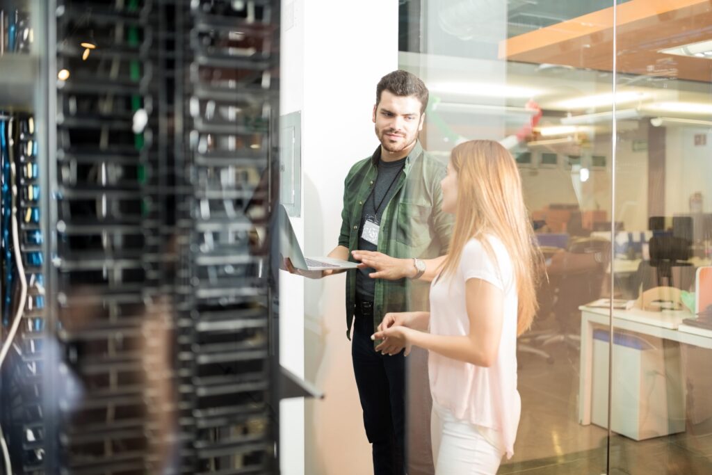 Two IT professionals discussing data in a server room, standing near server racks with a laptop