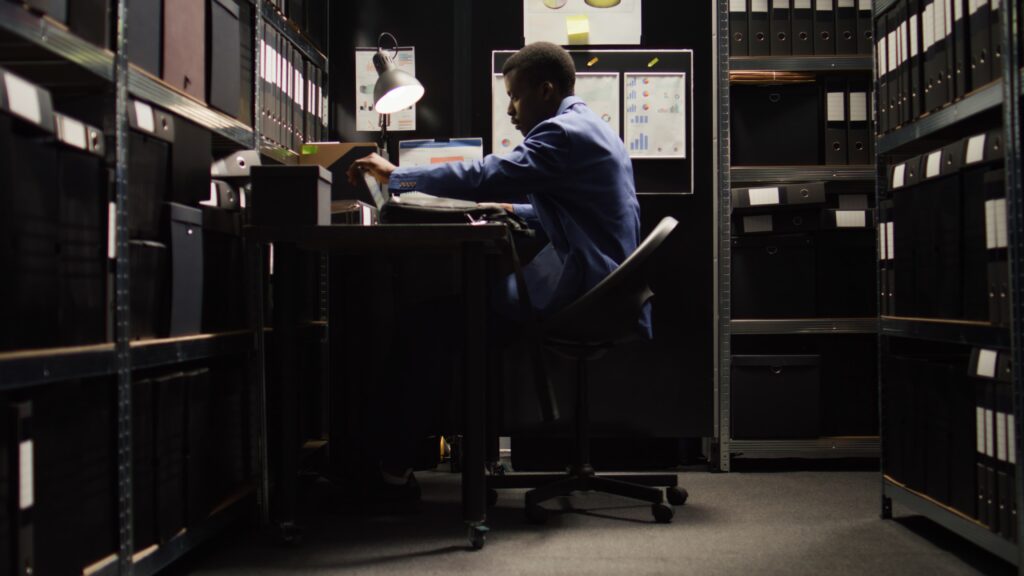 Private forensic investigator working on a laptop in a dimly lit office filled with files and case documents.